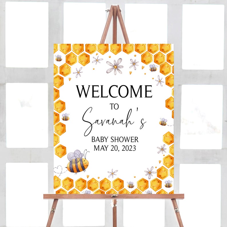 May include: Welcome to Savannah's Baby Shower sign featuring a honeycomb and bee design. The sign is white with black text and depicts a yellow and black bee, white flowers, and a honeycomb pattern.