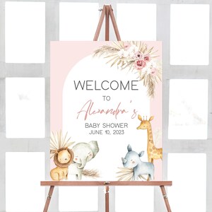 May include: A pink and white welcome sign for a baby shower with the text "Welcome to Alexandra's Baby Shower June 10, 2023". The sign features a watercolor illustration of a giraffe, elephant, lion, and rhino with a floral arrangement.