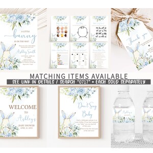 May include: A set of printable baby shower decorations featuring a blue and white bunny with floral accents. The set includes a welcome sign, a don't say baby sign, water bottle labels, a thank you tag, and a baby shower invitation. The text on the invitation reads "A little bunny is on the way!" and includes the name Ashley Smith.
