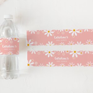 May include: Two pink and white daisy patterned labels with the text "Caroline's Baby Shower" printed on them. The labels are designed to wrap around a water bottle.