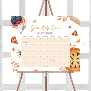 May include: A baby shower calendar on an easel. The calendar is light orange with a grid of dates for October and November. The text "Guess Baby Cara's BIRTH DATE" is at the top. Watercolor illustrations of pie slices, berries, and fall leaves decorate the edges.