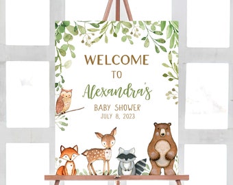 May include: Welcome to Alexandra's Baby Shower sign with a woodland theme. The sign features a green leafy border with a brown owl, fox, deer, raccoon, and bear. The text reads "Welcome to Alexandra's Baby Shower July 8, 2023".