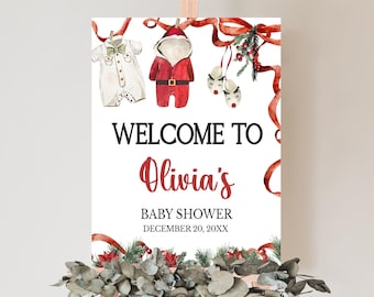 May include: A white sign on an easel with the words "WELCOME TO Olivia's BABY SHOWER DECEMBER 20, 20XX". The sign is decorated with watercolor illustrations of baby clothes and Christmas decorations. A garland of greenery and eucalyptus is at the base.