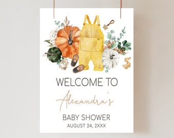 May include: A baby shower welcome sign featuring a watercolor illustration. The design includes a yellow jumpsuit, pumpkins, and the text "WELCOME TO Alexandra's BABY SHOWER AUGUST 24, 2XXX". The sign is suspended by a brown string.