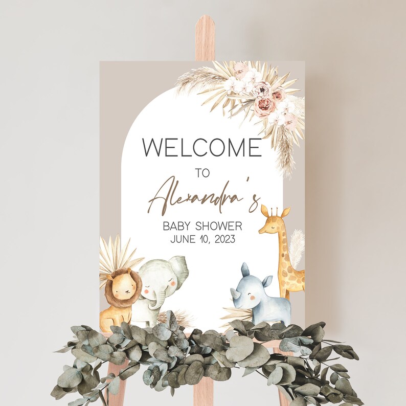 May include: Welcome to Alexandra's Baby Shower sign with a light brown background and a white circle with a floral design. The text "Welcome to Alexandra's Baby Shower June 10, 2023" is in brown text. The circle is surrounded by a light brown background with a floral design. There are three animals in the circle: a lion, an elephant, and a rhinoceros.