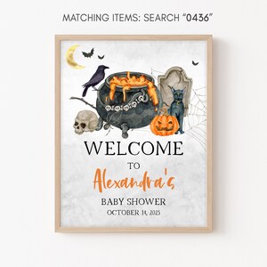 May include: A framed Halloween-themed baby shower sign. The watercolor design features a cauldron, black cat, pumpkin, skull, and the text "Welcome to Alexandra's Baby Shower October 14, 2023". The sign is in a light wood frame.