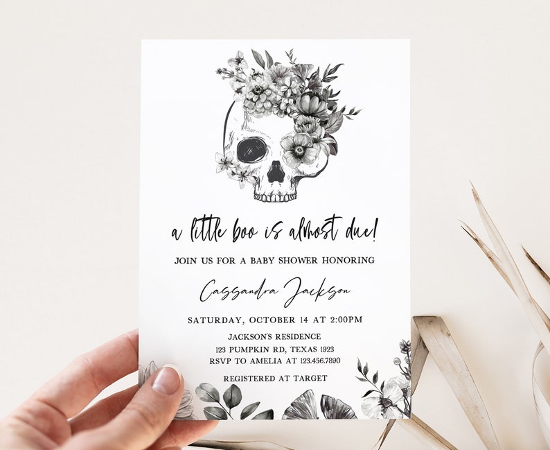 K&ouml;nnte beinhalten: Eine Babyparty-Einladung mit einem Totenkopf-Design, verziert mit floralen Elementen. Der Text lautet "a little boo is almost due!" und enth&auml;lt Veranstaltungsdetails wie Datum, Uhrzeit und Ort.