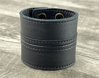Könnte beinhalten: Schwarzes Lederarmband mit zwei Druckknöpfen aus Messing. Das Armband hat zwei Reihen Nähte um die Mitte.