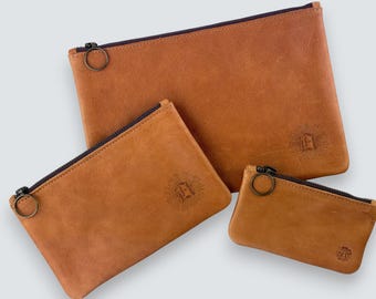 May include: Three tan leather pouches of varying sizes, each featuring a dark zipper with a circular pull tab. The two larger pouches display a stamped emblem of the letter E surrounded by sunburst lines, while the smallest has a smaller stamp.