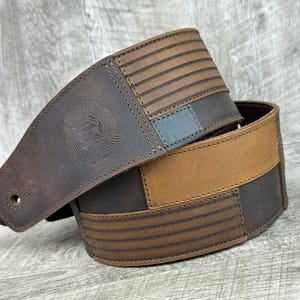 Może przedstawiać: Brązowy skórzany pasek do gitary z szytym wzorem. Pasek posiada logo w kształcie słońca i szereg poziomych linii. Pasek wykonany jest z różnych odcieni brązowej skóry.