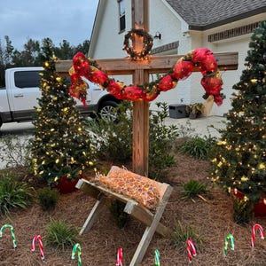 Puede incluir: Decoración navideña al aire libre con una cruz de madera adornada con una corona y una guirnalda roja. Un pesebre se encuentra debajo, flanqueado por dos árboles de Navidad iluminados y bastones de caramelo.