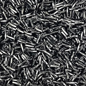 May include: A close-up of a pile of black glass seed beads. The beads are cylindrical with a hole through the middle. The beads are scattered, creating a textured surface.