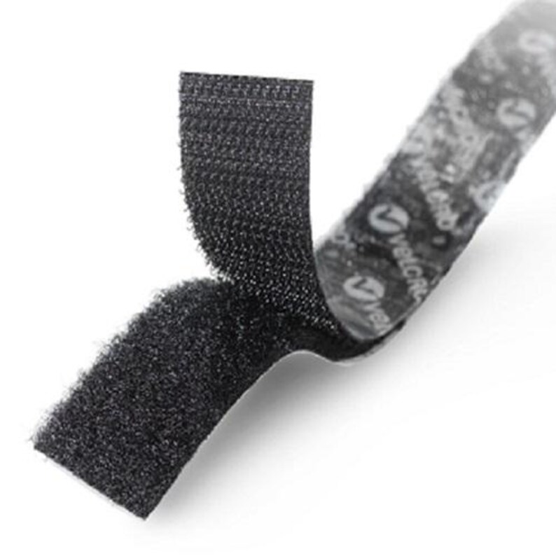 May include: A black hook and loop fastener strip with a textured surface. The strip is partially separated, revealing the adhesive backing which features the white Velcro brand logo printed repeatedly along the length of the adhesive side.