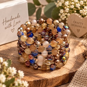 May include: A multi-strand wrap bracelet composed of various round beads in shades of brown, tan, clear, and blue, featuring a small silver-toned star charm. The bracelet is displayed alongside a white box with the text Handmade with Love.