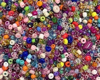 May include: A large collection of small, round glass beads in a wide variety of vibrant colors including red, blue, green, yellow, orange, purple, pink, white, and clear. The beads have a glossy finish and are scattered together in a dense pile.