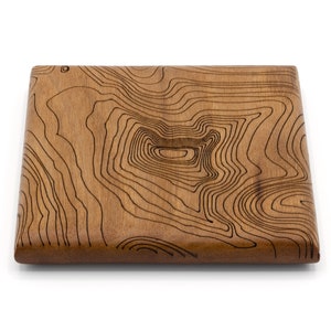 May include: A square wooden coaster with a brown wood grain finish and a laser-etched design of topographic lines.