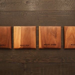May include: Four wooden squares with the names of mountains and their elevations engraved on them. The mountains are Mount Sneffels, Grays Peak, Mount Evans, and Wilson Peak.