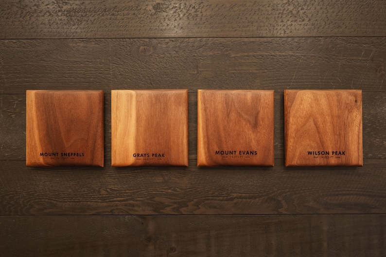 May include: Four wooden squares with the names of mountains and their elevations engraved on them. The mountains are Mount Sneffels, Grays Peak, Mount Evans, and Wilson Peak.
