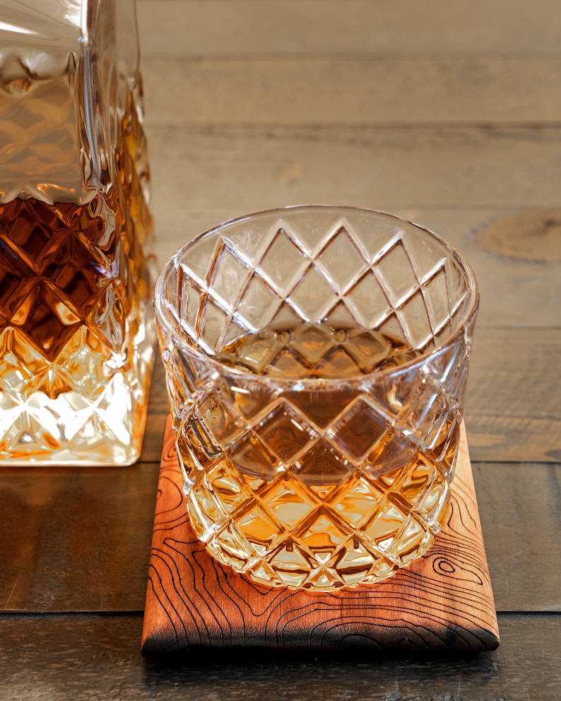 May include: A glass of amber-colored liquid sits on a wooden coaster. The glass has a diamond-shaped pattern and is filled with a clear liquid. The coaster is made of wood and has a natural grain.