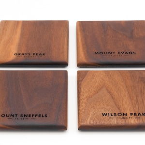 May include: Four wooden coasters with the names of mountains and their elevations in feet. The coasters are made of dark brown wood and have a natural grain pattern. The names of the mountains are "Grays Peak", "Mount Evans", "Mount Sneffels", and "Wilson Peak".