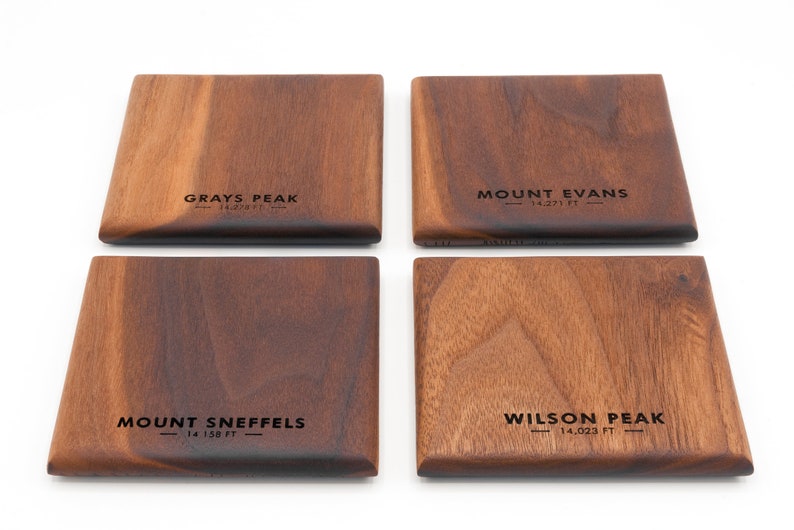 May include: Four wooden coasters with the names of mountains and their elevations in feet. The coasters are made of dark brown wood and have a natural grain pattern. The names of the mountains are "Grays Peak", "Mount Evans", "Mount Sneffels", and "Wilson Peak".
