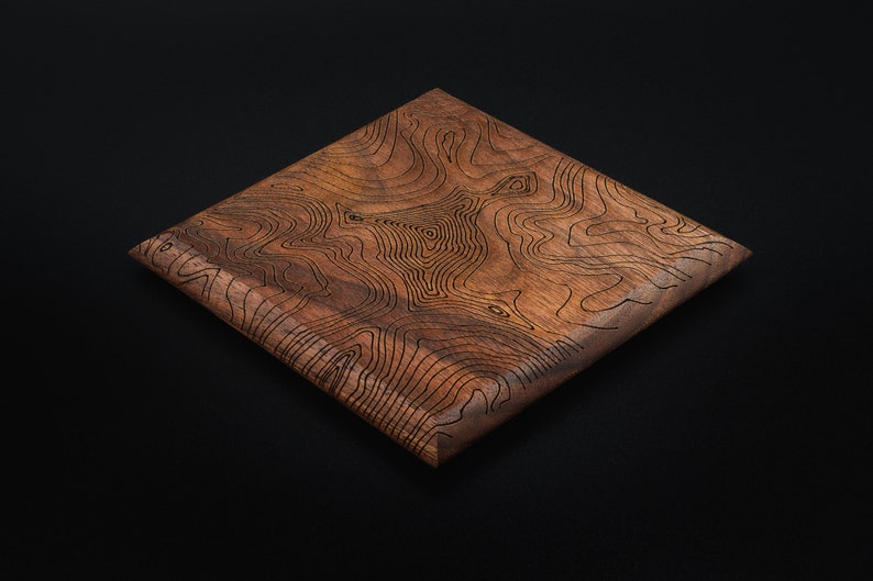 May include: A square wooden tray with a dark brown finish. The tray has a unique design of engraved lines that resemble a topographic map.