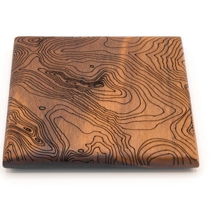 May include: A square wooden serving board with a dark brown wood grain and a black topographic map design.
