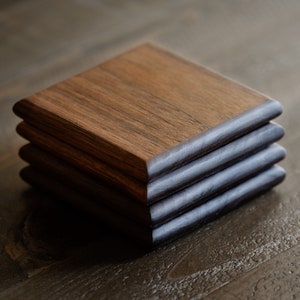 May include: A set of four square wooden coasters, stacked on top of each other. The coasters are made of dark wood with a natural grain pattern. The edges of the coasters are slightly rounded.