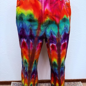 May include: Vibrant tie-dye jeans in a rainbow of colors, including red, orange, yellow, green, blue, and purple. The jeans have a classic five-pocket design with a button and zipper closure. The tie-dye pattern is vertically oriented.