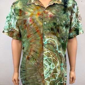 May include: Short-sleeved button-down shirt with a tie-dye pattern in shades of green, brown, and yellow. The shirt has two chest pockets and a collar. The tie-dye design features white accents.