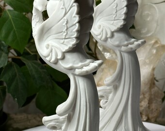 May include: A pair of white ceramic candle holders shaped like peacocks with intricate details. The holders are tall and slender, with the peacocks' tails extending out behind them.