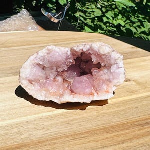 May include: A pale pink amethyst geode featuring a hollow center filled with clusters of translucent, light-colored crystalline structures. The mineral specimen has a rough, uneven outer surface and a jagged, crystalline interior cavity.