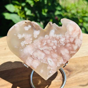 May include: A heart-shaped piece of polished flower agate stone featuring translucent light pink and cream-colored floral inclusions. The stone is displayed on a circular silver-toned metal stand, showcasing its smooth surface and natural crystalline patterns.