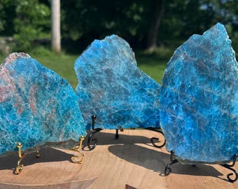 May include: Three raw, polished blue apatite crystal slabs displayed on decorative black metal stands with gold accents. The crystals have a translucent quality with visible internal patterns and some pinkish-red inclusions.