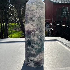 Puede incluir: Una gran punta de cristal de fluorita blanca, verde y morada. El cristal está pulido y tiene una superficie lisa y brillante. Está sobre una superficie blanca.