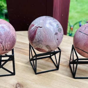 May include: Three polished spherical stones with pink, white, and grey marbled patterns, each resting on a black geometric wire stand. The spheres display natural veining and color variations, showcasing smooth surfaces and distinct mineral textures.