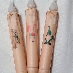May include: Three battery-operated LED candles with a rustic brown finish. The candles feature a bunny, a gnome, and a bunny with an Easter egg.