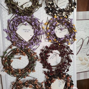 May include: Assortment of eight wreaths made of brown twigs with various colored berry accents. The wreaths are arranged in a circle on a white surface. The wreaths are all the same size and are approximately 10 inches in diameter. The berries are in the following colors: green, white, black, purple, yellow, red, brown, and orange.