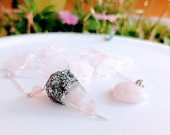 May include: A crystal pendulum with a silver chain and a pink gemstone. The pendulum has a cone-shaped crystal with a silver-colored top. A small, rounded pink gemstone is attached to the chain.