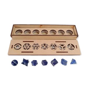 Puede incluir: Un juego de caja de madera con siete formas geométricas y siete piedras de lapislázuli azul. La caja tiene una tapa con diseños geométricos recortados. Las piedras tienen varias formas, incluyendo esferas, pirámides y cubos.