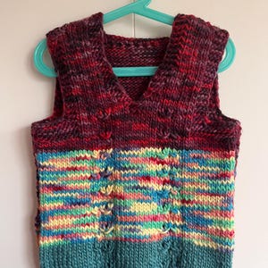 Può includere: Un gilet lavorato a mano a maglia con scollo a V, caratterizzato da un design a blocchi di colore. La sezione superiore è di un bordeaux intenso con macchie rosse, mentre la parte inferiore mostra vibranti strisce arcobaleno e verde acqua. Il gilet è esposto su una gruccia turchese.