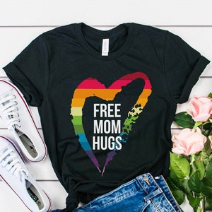 Op de afbeelding: Een zwart t-shirt met korte mouwen, een regenboogkleurig hartmotief en de witte tekst FREE MOM HUGS, gecombineerd met een blauwe spijkerbroek en witte canvas sneakers. Het ontwerp bevat een hartvorm met horizontale gekleurde strepen.