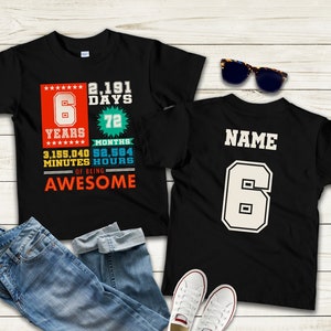 May include: Two black t-shirts with a white graphic design. The front shirt has a colourful design with the text "6 Years 2,191 Days 72 Months 3,155,040 Minutes 52,584 Hours of Being Awesome". The back shirt has the text "NAME" and the number "6" in white.