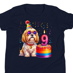 May include: Navy blue t-shirt featuring a cartoon dog wearing sunglasses and a party hat. The dog is next to a rainbow-colored cake with a lit candle shaped like the number 9. Colorful confetti is scattered around the design.