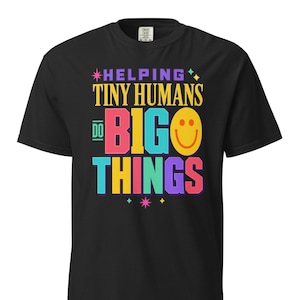Peut inclure: Un t-shirt noir à manches courtes avec le texte coloré HELPING TINY HUMANS DO BIG THINGS et un visage souriant jaune remplaçant la lettre O dans BIG. Le motif comprend de petites étoiles décoratives en rose, bleu sarcelle et jaune.