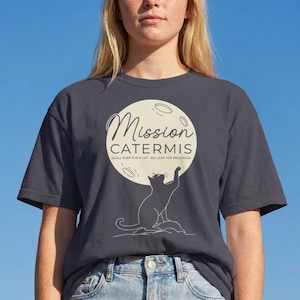 May include: Dark gray t-shirt featuring a cat silhouette reaching for a moon graphic. The graphic includes the text "Mission Catermis" and the tagline "Small Paws for a Cat - Big Leap for Meowning". The shirt is a casual, everyday wear item.