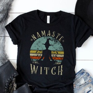 Può includere: T-shirt nero con la scritta "NAMASTE WITCH" in un design circolare con la silhouette di una strega. La maglietta è abbinata a pantaloncini di jeans, stivali neri e un cappello nero. Decorazioni di Halloween con pipistrelli e ragnatele.