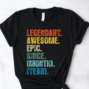 May include: Black t-shirt featuring the text "LEGENDARY. AWESOME. EPIC. SINCE. (MONTH). (YEAR)." in a retro, multi-coloured font. The shirt has a crew neck and is made of a soft material.