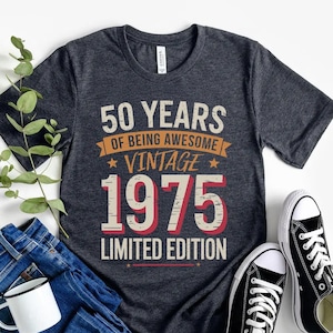 May include: A dark gray t-shirt with the text "50 Years of Being Awesome Vintage 1975 Limited Edition" in red, white, and yellow lettering. The shirt is laid flat on a white surface with a pair of black sneakers and blue jeans.