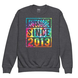May include: A dark grey sweatshirt featuring the text "AWESOME SINCE 2013" in a vibrant tie-dye pattern. The text is enclosed within a rectangular border. The sweatshirt has a crew neck and long sleeves.